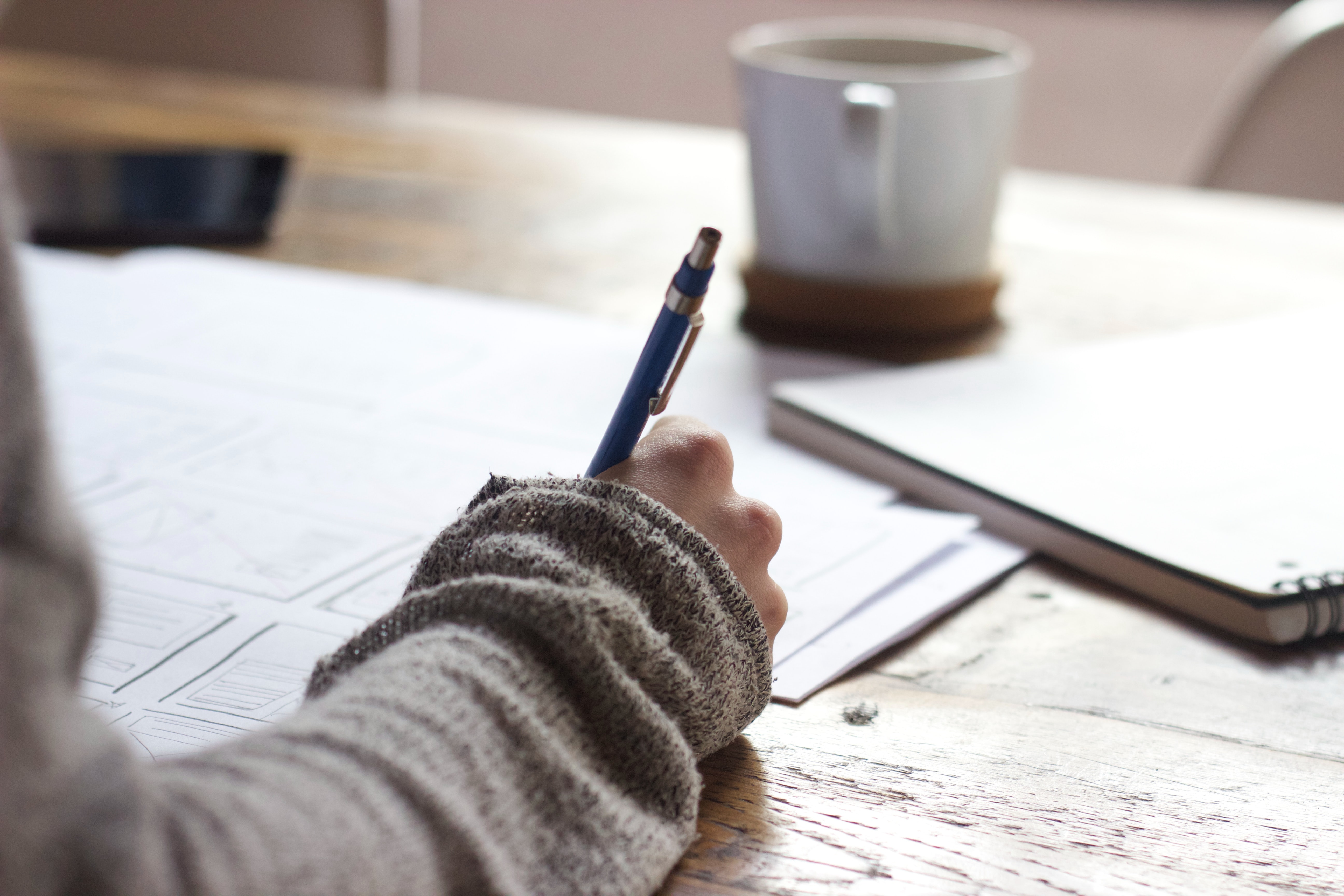 Picture of a female student wearing a long sleeved sweater writing on a paper with a notebook and cup of coffee next to her.